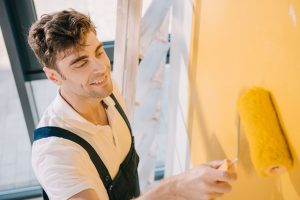 handsome-young-painter-smiling-while-painting-wall-CVNVD59.jpg